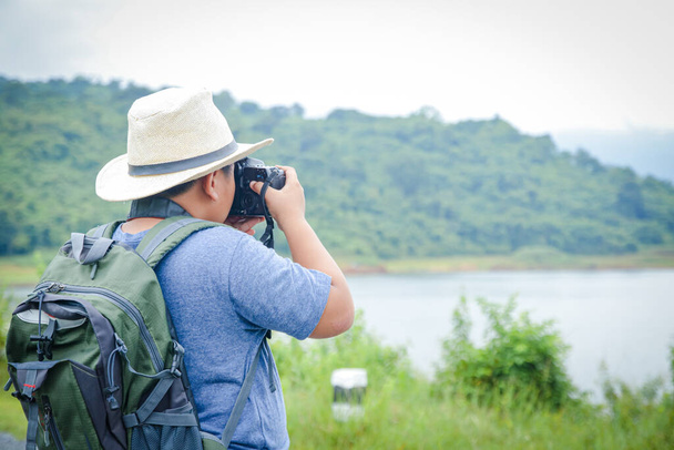 Um pequeno menino asiático usando um chapéu gosta de viajar para a natureza. Segure a câmera para tirar uma foto da paisagem - Foto, Imagem