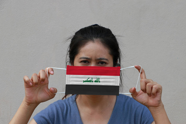 Жінка з іракським прапором на гігієнічній масці в руці і підняла переднє обличчя на бежевому тлі. Крихітні частки або вірус корони або ковидки 19 захисту. Концепція боротьби з хворобами. - Фото, зображення