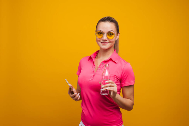 Cute blonde in sunglasses and t-shirt, drinking water from a bottle, portrait on a yellow background - Photo, image