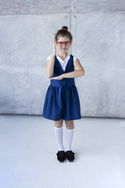 Fröhliches Teenie-Mädchen mit roter Brille, weißem T-Shirt und Schuluniform posiert im Studio und blickt in die Kamera, Betonhintergrund. Zurück zu den Schulinhalten. - Foto, Bild