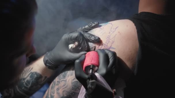 Nagranie tatuażysty robiącego tatuaż na ramieniu mężczyzny w salonie - Materiał filmowy, wideo
