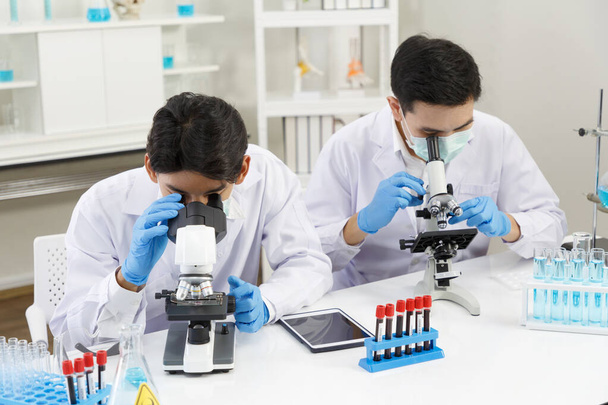 Dos científicos masculinos usan mascarilla facial que trabaja en el laboratorio mientras verifican la solución por microscopio. SARS-CoV-2, Covid-19 TEMA. - Foto, Imagen