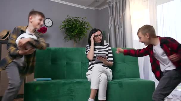 Attractive concentrated busy young woman in glasses working with tablet pc and feeling headache becouse her noisy playful teen sons running around the couch on that she sits - Záběry, video