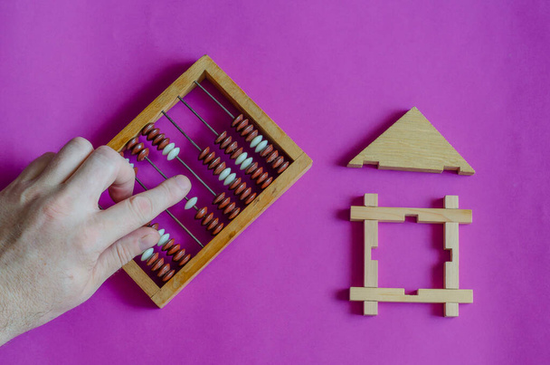 Male hand calculates an abacus next to toy wooden house on lilac background. The concept of mortgage or lending secured by real estate. View from above. - Photo, Image