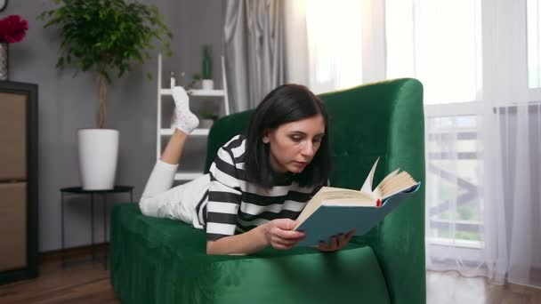 Education concept where good-looking concentrated young woman with dark hair lying on comfortable sofa and reading book - Imágenes, Vídeo