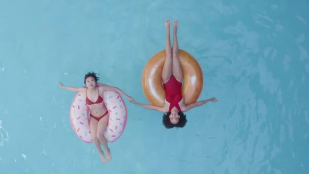 Two people asian and afro american women in red swimsuit relaxing refreshing resting in inflatable swimming tube ring circle pink donut in pool home hotel beach tourist complex, top view, full length - Footage, Video