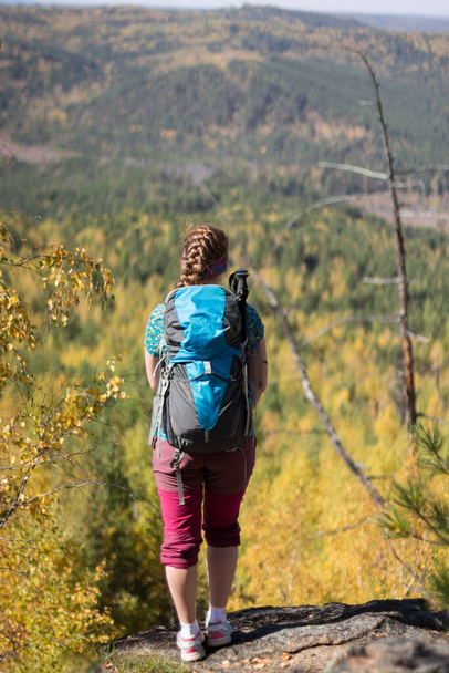 Red-haired girl standing with a backpack on a rock on a background of autumn forest - Foto, Imagen