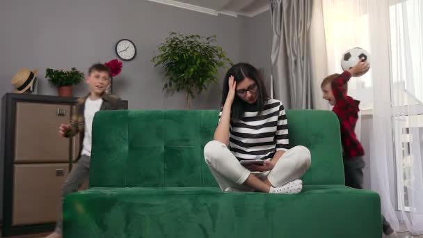Attractive tired concentrated young woman sitting on the couch at home and feeling headache from the noise that creating her two playful loud sons near her - Záběry, video