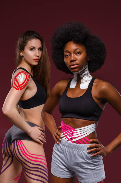 Kinesiology taping. Two young female athletes on red background with kinesiology tape on neck,shoulder, khee, tummy and hips. Fat lose, cellulite removal, sport physical therapy,recovery concept - Φωτογραφία, εικόνα