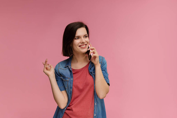 Young people working with mobile devices. Portrait of cute happy brunette woman wearing t-shirt and jeans shirt talking on mobile phone and smiling isolated over pink background. Copy space - Photo, image