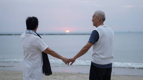 Asian elder senior couple holding hands looking sunset sea ocean together happy retirement life - Foto, imagen