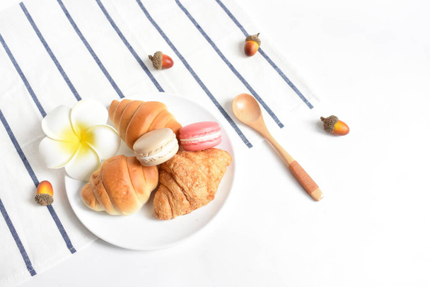 Flat lay, bovenaanzicht, eten en drinken in witte achtergrond, gezonde ontbijtmaaltijd met croissants, Franse Macaron, aardbei, gedroogde eikel, lepel, kers, witte Fragipane bloem - Foto, afbeelding