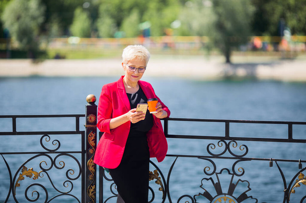 beautiful elderly woman with short hair and glasses with a phone and a cup of coffee in her hands - Fotó, kép