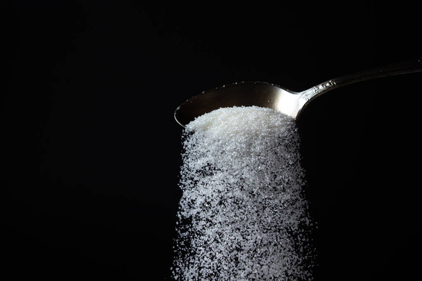 黒の背景にスプーンから砂糖を注ぐ。過剰な砂糖摂取。甘い結晶 - 写真・画像