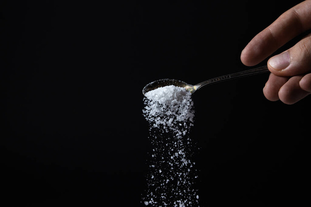 Соль наливают из ложки на черном фоне. Чрезмерное потребление соли. Грубая соль - Фото, изображение