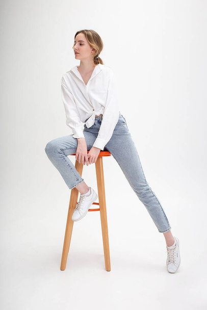 young pensive caucasian woman posing in shirt and blue jeans, sitting on stool - Photo, Image