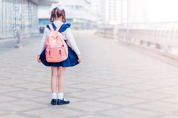Una niña en un vestido de escuela azul con una mochila rosa en la espalda, coletas y arcos se coloca con su espalda a la cámara. Fotografía horizontal - Foto, Imagen