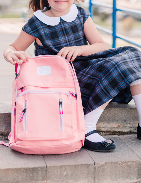 Una niña en un vestido de escuela azul, medias blancas y zapatos se sienta en los escalones con una mochila rosa. Foto vertical - Foto, imagen