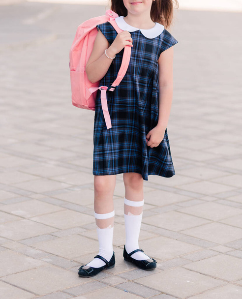 Una chica con un vestido de escuela azul y una mochila rosa en la espalda. Foto vertical - Foto, Imagen
