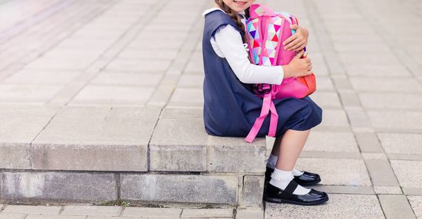 Dívka v modrých školních šatech, bílých ponožkách a botách sedí na schodech s růžovým batohem. Vodorovná fotka. Kopírovat prostor - Fotografie, Obrázek