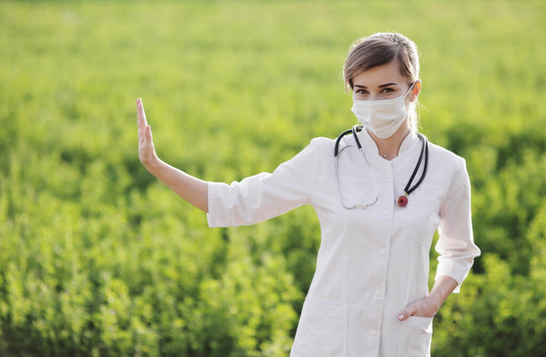 Uma médica com uma máscara facial médica que mostra um gesto de parar a pandemia. Médica ou enfermeira confiante usando uma máscara protetora facial no fundo de grama verde. Coronavirus Covid19. Rapariga, mulher
 - Foto, Imagem