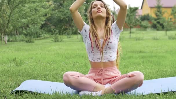 Young beautiful woman with dreadlocks is doing yoga sitting in lotus position outdoors during morning on green grass of her backyard - Footage, Video