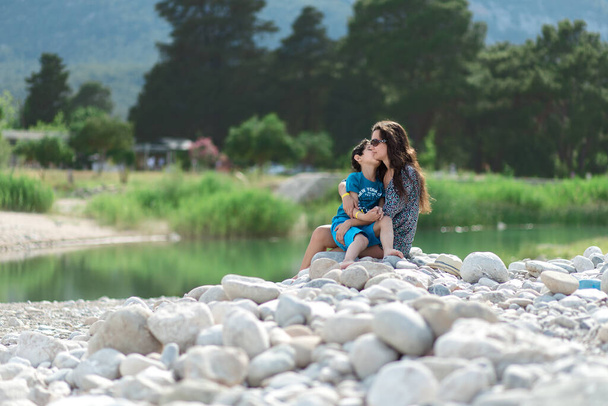 Kobieta i chłopiec siedzą w pobliżu rzeki lub jeziora. Matka i syn. Dzień Matki. Rodzina - Zdjęcie, obraz
