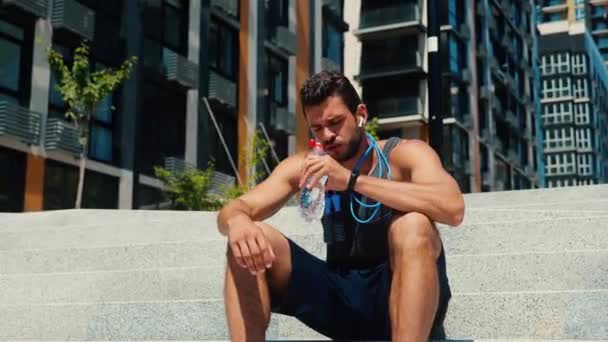 Young man exercising outside. Tired exhausted guy sitting on steps on street and drink water. Jumping rope is hanging around his neck. After running or jogging outside. - Footage, Video