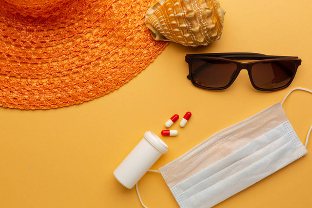 保護面マスク、ビーチでのパンデミック休暇の概念とオレンジの背景にサングラスや帽子。コロナウイルス感染時間の観光と休暇. - 写真・画像