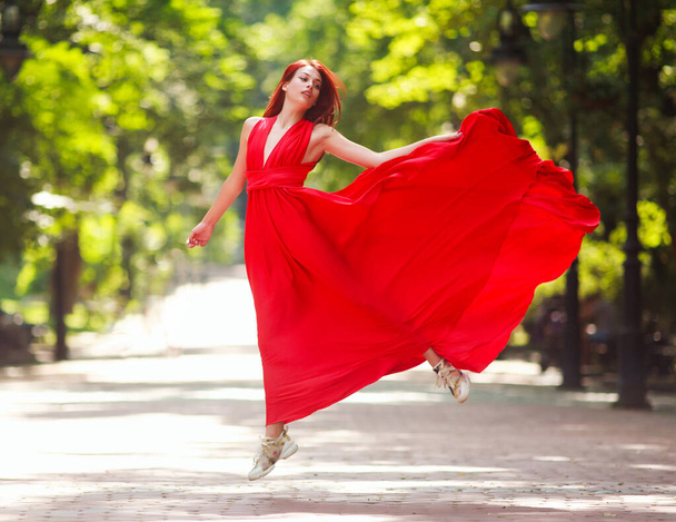 Young woman in a luxury long red dress dancing, jumping in city park - Φωτογραφία, εικόνα