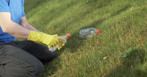 Чоловік в жовтих рукавичках, видаляючи сміття з зеленої трави, скручує і стискає пластикові пляшки для подальшого сортування
. - Кадри, відео