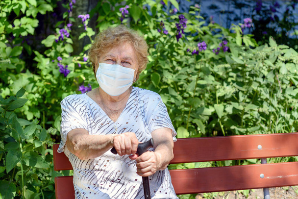 Una donna anziana in una maschera protettiva medica N95 è seduta su una panchina del parco. - Foto, immagini