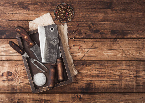 Vintage hatchets for meat in wooden box with salt and pepper on wooden background with linen towel and fork and knife. Space for text - 写真・画像