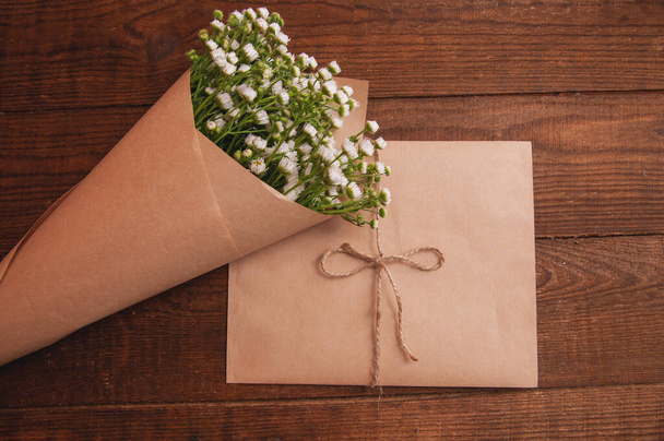 envelope made of craft paper, next to a bouquet of chamomile flowers, which lies on a wooden table - Фото, изображение