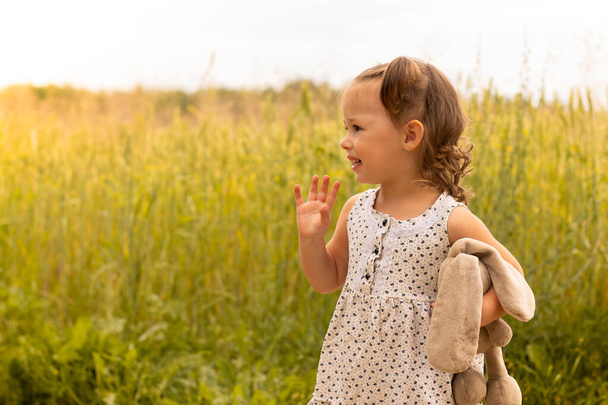 Kis aranyos lány 1-3 egy plüss nyúl egy könnyű ruha integetett a kezét a területen tüskés rozs nyáron - Fotó, kép