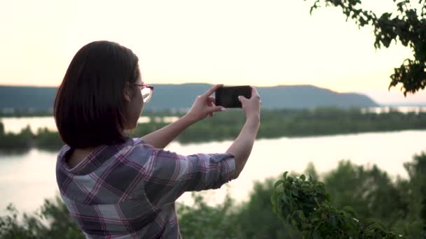 Mladá žena stojí na kopci na pozadí řeky a hor a fotografuje krajinu. Dívka fotí v přírodě. - Záběry, video