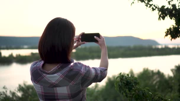 Une jeune femme se tient sur une colline sur le fond de la rivière et des montagnes et photographie le paysage. La fille prend des photos dans la nature. - Séquence, vidéo