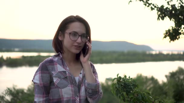 Een jonge vrouw staat op een heuvel tegen de achtergrond van de rivier en de bergen en spreekt aan de telefoon. Het meisje is aan de telefoon in de natuur close-up. - Video