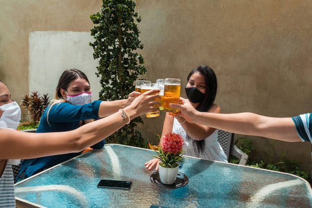 Amigos enmascarados bebiendo cerveza en casa, divirtiéndose. Parque de máscaras covid - Foto, Imagen