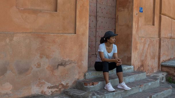 Γυναίκα κάθεται στα σκαλιά μπροστά από την πόρτα με καπέλο και αθλητικά παπούτσια στην Μπολόνια Ιταλία κοιτάζοντας προς τα αριστερά της - Φωτογραφία, εικόνα