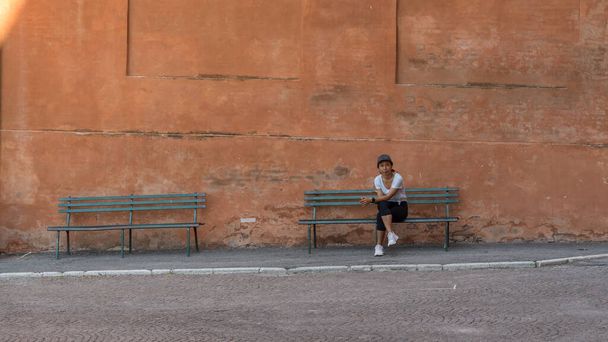 Mujer con sombrero, camiseta, pantalones negros y zapatillas de deporte sentada en el banco del parque con las piernas cruzadas en Bolonia con otro banco del parque a su derecha - Foto, Imagen