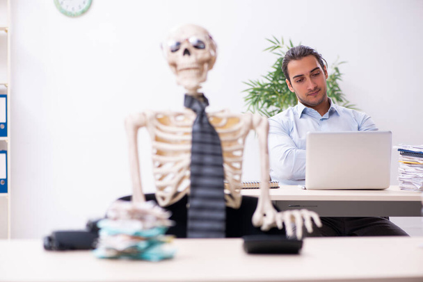 Νεαρός άνδρας υπάλληλος και σκελετός στο γραφείο - Φωτογραφία, εικόνα