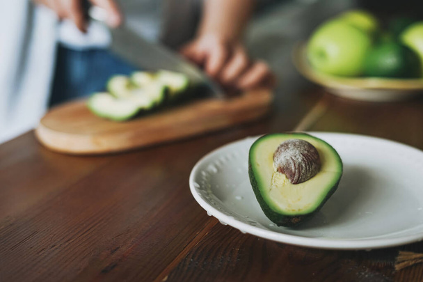 Female hands cut avocado on wooden working surface in kitchen at home - 写真・画像