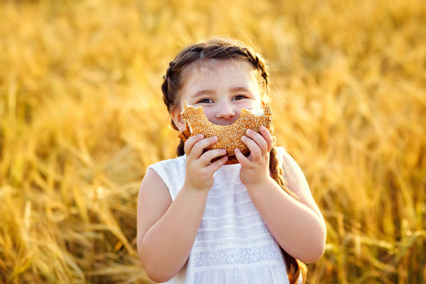Een lachend klein meisje in een witte zonnejurk en twee staarten op een tarweveld met een bagel in de open lucht. Hij zet een bagel op zijn mond in de vorm van een glimlach. - Foto, afbeelding