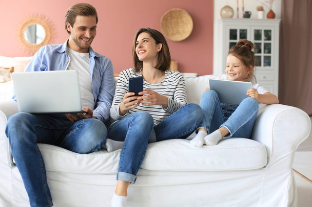 Father, mother and daughter using electronic devices sitting on sofa at living room - Photo, image