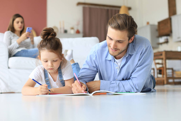 Happy father smilling daughter lying on warm floor enjoying creative activity, drawing pencils coloring pictures in albums, mother resting on couch, family spend free time together - Foto, imagen