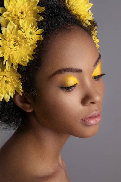 Young sensual african american woman with artistic make-up and gerbera in hair isolated on grey background - Photo, Image