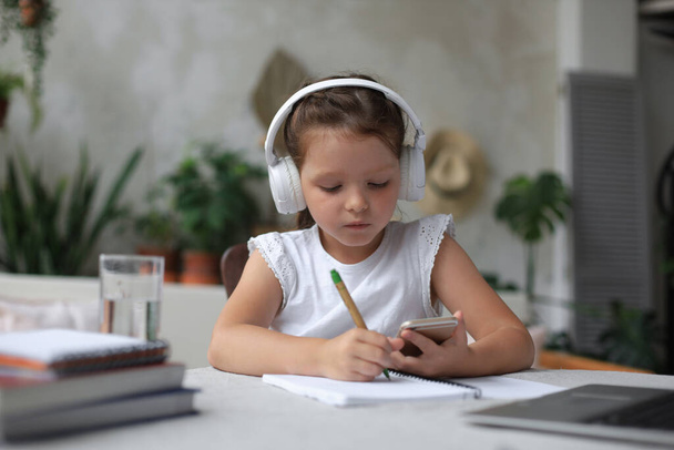 Smart little girl in headphones do homework online class on smartphone, small child in earphones study on Internet, have web conference or lesson during quarantine - Photo, Image
