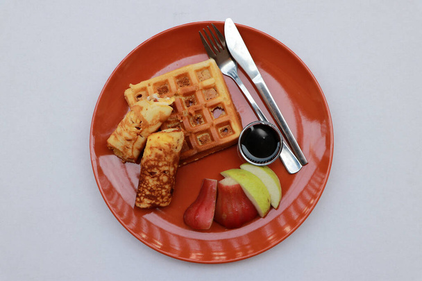 Breakfast in the dark orange color of round plate on the white floor. Dough roll on the waffle and fruit rose apple, guava with knife and fork. - Fotó, kép