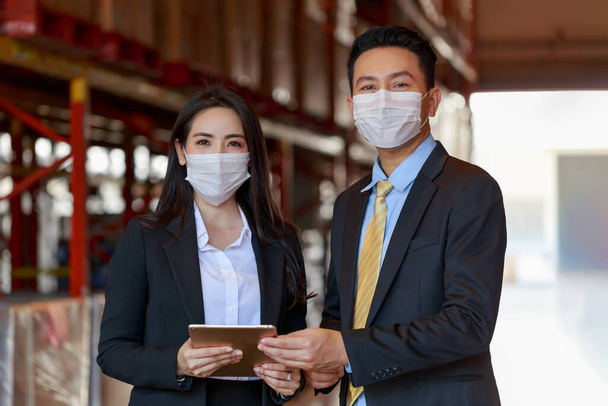 Retrato del atractivo hombre de negocios asiático y la mujer inversora con máscara facial protectora en el almacén, nuevo normal después de la pandemia del virus Covid-19 Corona. - Foto, imagen